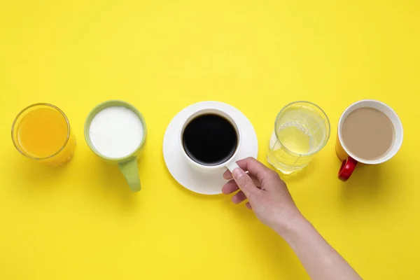La mano femenina sostiene la taza con café negro de un conjunto de bebidas jugo de naranja, café con leche, agua justa, yogur sobre un fondo amarillo. Piso tendido, vista superior — Foto de Stock