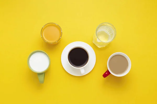 Grupo de bebidas en tazas multicolores, café negro, café con leche, yogur, agua, jugo de naranja sobre fondo amarillo. Piso tendido, vista superior — Foto de Stock