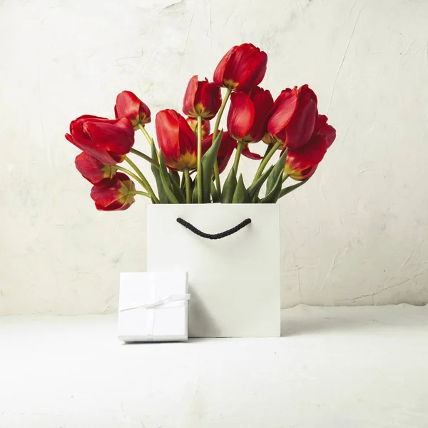 White Gift bag, small white gift box and bouquet of red tulips on a light stone background. Concept Offers an engagement or marriage