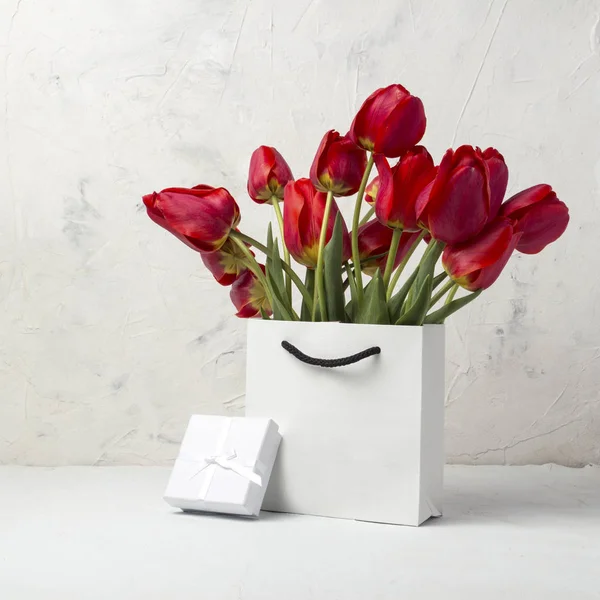 White Gift Bag, small white gift box, petals and bouquet of red tulips on a light stone background. Concept Offer a gift or an engagement, marriage