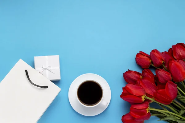 White Gift Bag, a small white gift box, a white cup with black coffee and a bouquet of red tulips on a blue background. Concept Offers an engagement or marriage, shopping