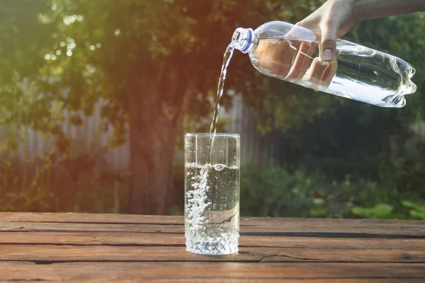 Female hand pours water from a plastic bottle into a glass on a wooden table in a spring garden
