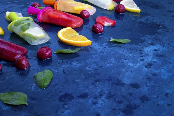 Homemade bright fruit popsicle with strawberry, cherry, lemon, orange, lemon and mint flavor and fresh fruit for ice cream on a dark blue background