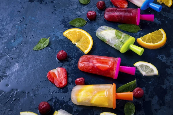 Multicolored bright fruit popsicle with strawberry, cherry, lemon, orange, lemon and mint and slices fresh fruit on a dark blue background