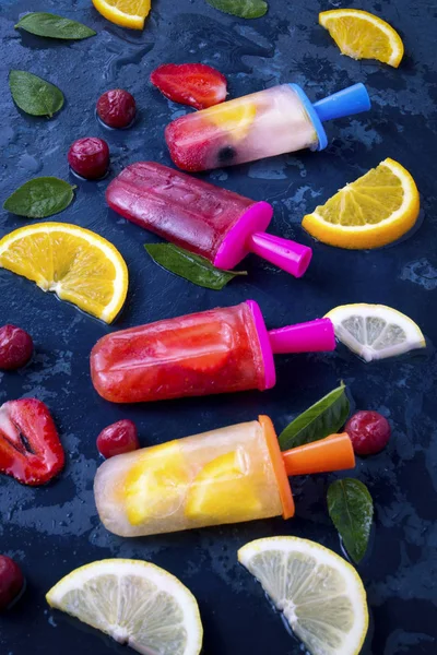 Multicolored bright fruit popsicle with strawberry, cherry, lemon, orange, lemon and mint and slices fresh fruit on a dark blue background