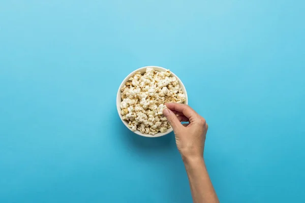 Mão feminina tomando pipocas de uma xícara branca em um fundo azul. Conceito assistindo a um filme com pipocas, atividades de lazer para comer pipocas. Deitado plano, vista superior — Fotografia de Stock