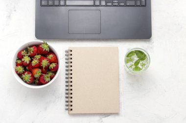 Çalışma alanı. Laptop, defter, beyaz bir kase çilek ve serinletici bir içecek buz ve nane ile. Üstten görünüm, düz yatıyordu.