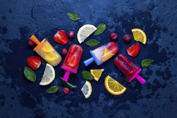 Homemade bright fruit popsicle with strawberry, cherry, lemon, orange, lemon and mint flavor and fresh fruit for ice cream on a dark blue background. Flat lay, top view
