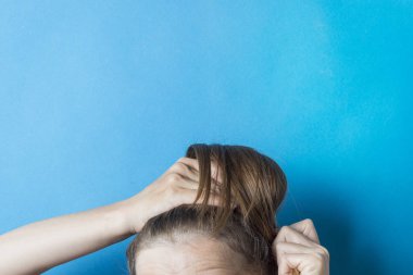 Kız, kafasındaki dağınık saç kuyruğunu mavi arka plana doğru düzeltiyor. Modern hızlı saç stili. Lastik bantla bağlanmış saçlar.