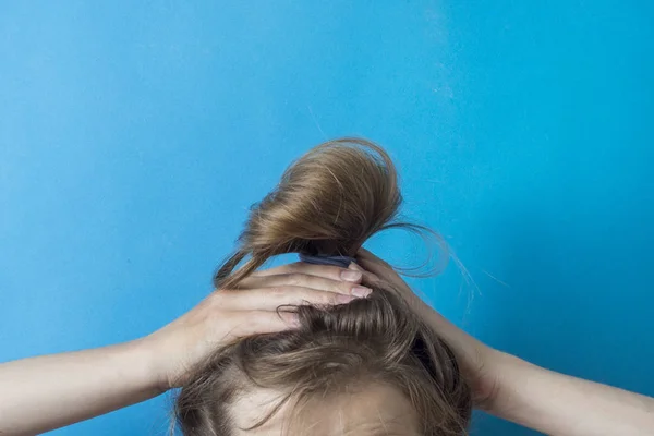 Girl straightens the disheveled tail of hair on her head, against a blue background. Modern fast hairstyle. Hair tied with elastic band