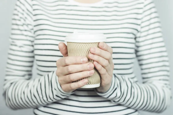 Una chica con una camiseta a rayas sostiene una taza de papel con café o té frente a ella. Concepto de comida para llevar, cafetería . — Foto de Stock