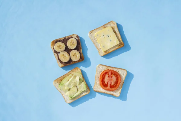 Σάντουιτς με τυρί, ντομάτα, μπανάνα και αβοκάντο σε μπλε φόντο. Η ιδέα της υγιεινής διατροφής, το πρωινό στο ξενοδοχείο, η δίαιτα. Φυσικός φωτισμός, σκληρό φως. Επίπεδη όψη, κορυφή θέα. — Φωτογραφία Αρχείου