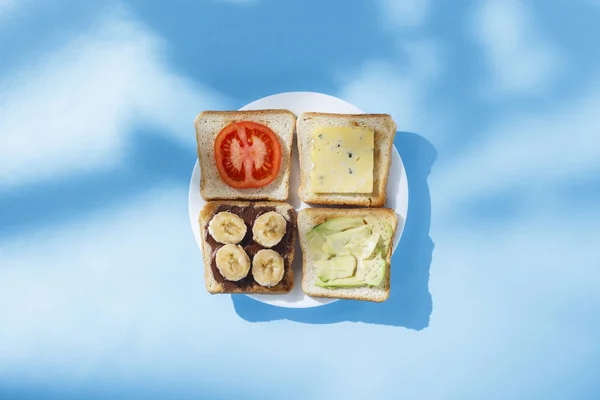 Sandwiches mit Käse, Tomate, Banane und Avocado auf weißem Teller, blauem Hintergrund. Konzept der gesunden Ernährung, Frühstück im Hotel, Ernährung. natürliches Licht, hartes Licht. flache Lage, Draufsicht. — Stockfoto