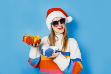 Noel Baba şapkalı genç bir kız mavi bir arka plan üzerinde hediye kutuları tutuyor. Yeni yıl ve Noel kavramı, kış tatilleri için hediyeler, satış alışveriş.