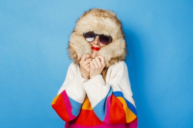 Kürk şapkalı mutlu yüzlü, gözlüklü ve mavi arka planda süveter giyen bir kız. Kış, kış tatilleri ve satış konsepti.