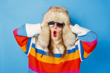 Kürk şapkalı mutlu yüzlü, gözlüklü ve mavi arka planda süveter giyen bir kız. Kış, kış tatilleri ve satış konsepti.