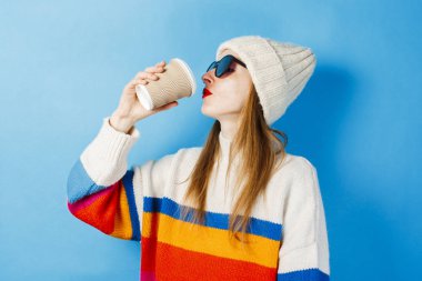 Gözlüklü ve şapkalı genç bir kız, mavi arka plandaki kağıt bardaktan kahve ya da çay içiyor. Konsept kahve dükkanı, kış, hipster.