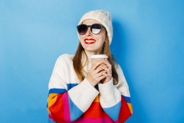 Gözlüklü ve şapkalı genç bir kız gülümsüyor ve mavi bir arka plan aleyhine çay veya kahve ile bir kağıt fincan tutuyor. Konsept kahve dükkanı, kış, hipster.