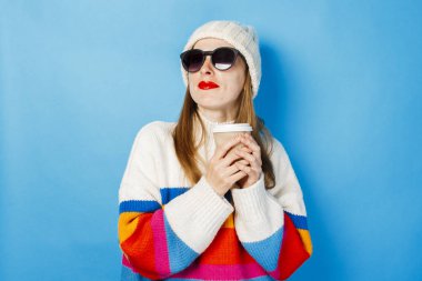 Gözlüklü ve şapkalı genç bir kız gülümsüyor ve mavi bir arka plan aleyhine çay veya kahve ile bir kağıt fincan tutuyor. Konsept kahve dükkanı, kış, hipster.