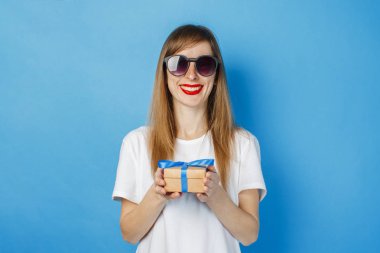 Gözlüklü ve beyaz tişörtlü genç bir kız mavi arka planda bir hediye kutusu tutuyor. Doğum günü veya tatil hediyesi konsepti.