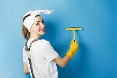 Bir gülümseme ve kafasına bir mendil ile genç bir kız mavi bir arka plan üzerinde bir shkrebk ile kir siliyor. Temizlik Konsepti ve Temizlik Hizmeti.