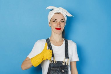 Başında eşarp ve sarı lastik eldivenli genç bir kız mavi arka planda olduğu gibi baş parmak hareketi yapıyor. Temizlik ve temizlik hizmeti kavramı, yüksek kalite.