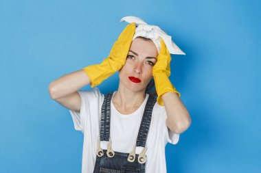Yorgun ve üzgün bir yüzü ve kafasında mendili olan genç bir kız, mavi arka planda başını tutuyor. Temizlik ve temizlik hizmeti kavramı, yorgunluk, sıkı çalışma, zor program.