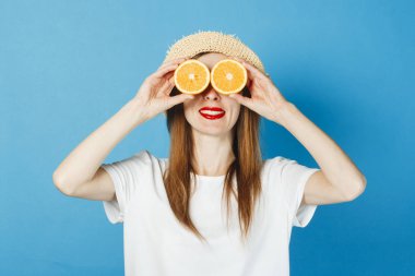 Genç bir kız gözlük, beyaz bir t-shirt ve hasır şapka, mavi bir arka plan gibi gözlerinin yanında portakal tutar. Konsept tatil, turistik gezi, yaz ve tropik.