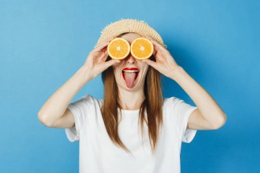 Genç bir kız gözlük, beyaz bir t-shirt ve hasır şapka, mavi bir arka plan gibi gözlerinin yanında portakal tutar. Konsept tatil, turistik gezi, yaz ve tropik.