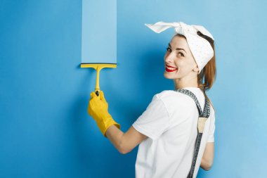 Bir gülümseme ve kafasına bir mendil ile genç bir kız mavi bir arka plan üzerinde bir shkrebk ile kir siliyor. Temizlik Konsepti ve Temizlik Hizmeti.
