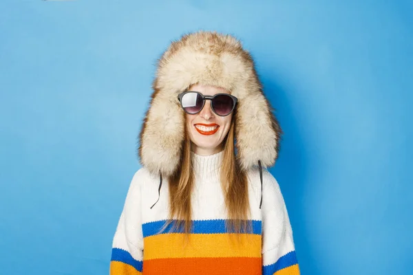 파란색 배경에 모피 모자, 안경과 스웨터에 행복한 얼굴을 가진 소녀. 겨울, 겨울 휴가 및 판매의 개념. — 스톡 사진