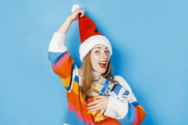 Una joven con un sombrero de Santa Claus sostiene cajas de regalo sobre un fondo azul. El concepto de Año Nuevo y Navidad, regalos para las vacaciones de invierno, compras en las ventas . — Foto de Stock