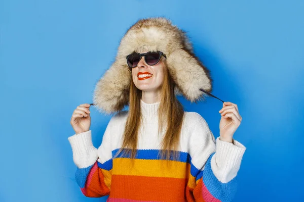 Uma menina com um rosto feliz em um chapéu de pele, óculos e uma camisola em um fundo azul. O conceito de inverno, férias de inverno e vendas . — Fotografia de Stock