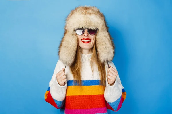 파란색 배경에 모피 모자, 안경과 스웨터에 행복한 얼굴을 가진 소녀. 겨울, 겨울 휴가 및 판매의 개념. — 스톡 사진