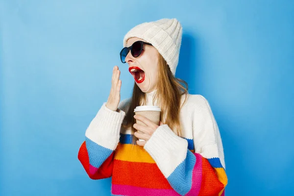 Una joven en vasos y un sombrero bosteza y sostiene una taza de papel con té o café sobre un fondo azul. Concepto cafetería, invierno, café para la alegría . — Foto de Stock