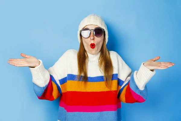 흰색 모자, 안경, 파란색 배경에 스웨터에 놀란 얼굴을 가진 어린 소녀. 겨울의 개념, 밀리, 힙스터. — 스톡 사진