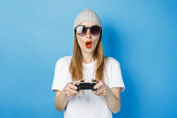A girl in glasses, a hat and a white T-shirt is holding a gamepad on a blue background. Concept game on the console, cyber sports, entertainment. — Stock Photo, Image