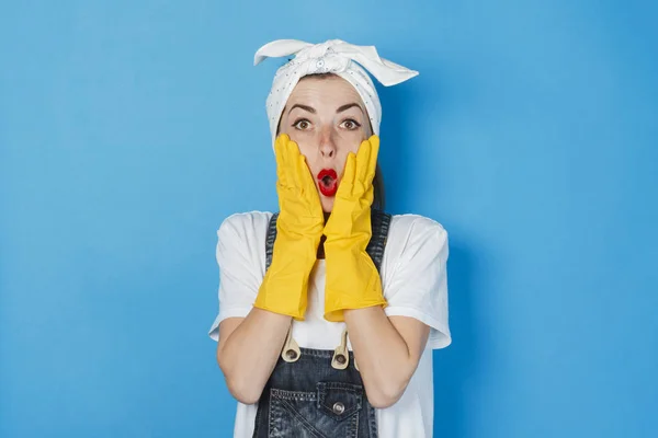 Een jong meisje met een verrast gezicht en een sjaal op haar hoofd op een blauwe achtergrond. Het concept van het schoonmaken en schoonmaken service, hoge kwaliteit. Schok, veel werk. — Stockfoto
