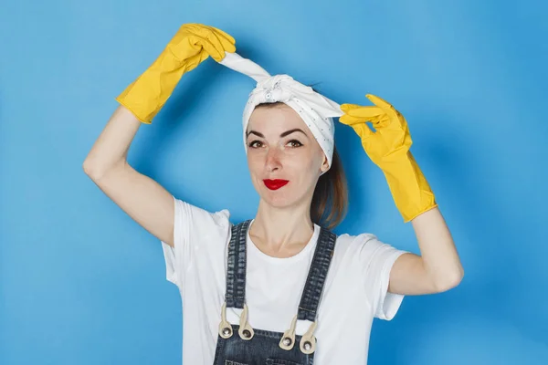 A young girl with a scarf on her head, yellow rubber gloves, tying a bandana on her head against a blue background. The concept of cleaning and cleaning service, high quality.