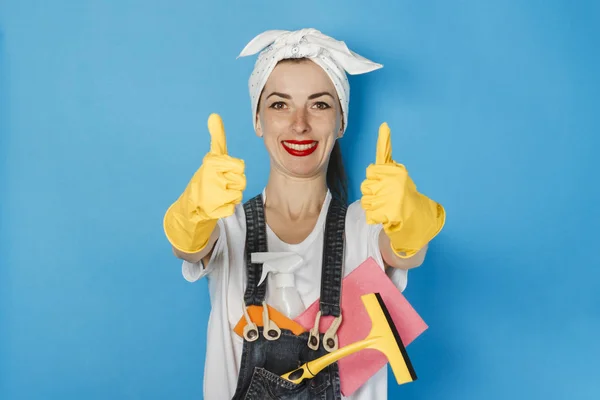 Chica joven con una bufanda en la cabeza, guantes de goma amarilla y artículos de limpieza que muestran un gesto hacia arriba pulgares, super, como en un fondo azul. Concepto de servicio de limpieza y limpieza, alta calidad . — Foto de Stock