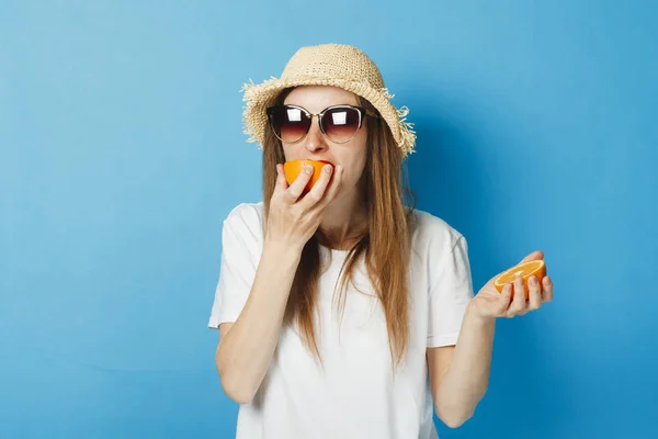 Молодая девушка в очках и соломенной шляпе кусает оранжевый, синий фон. Концепция отдыха, туристическая поездка, лето и тропики . — стоковое фото