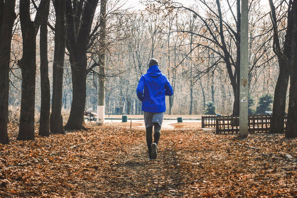 A man in a blue jacket makes a run in an autumn or spring park. Jogging concept, workout outdoor.