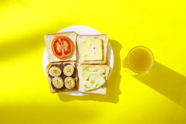 Sandwiches mit Banane, Tomate, Avocado, Blauschimmelkäse auf weißem Teller und einem Glas mit Orangensaft auf gelbem Hintergrund. Konzept eines gesunden Frühstücks, Vitamine, Ernährung. flache Lage, Draufsicht. — Stockfoto