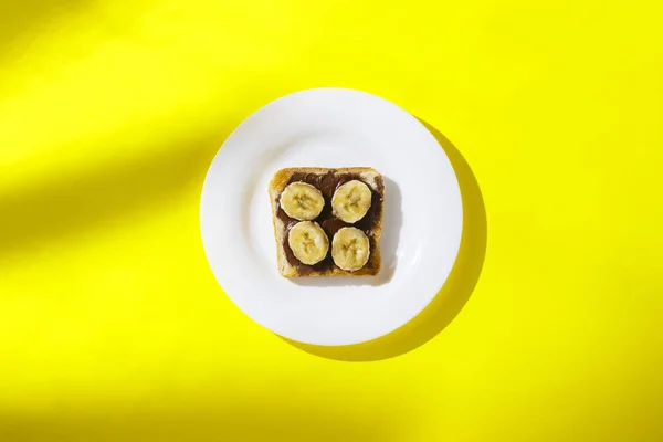 Σάντουιτς με μπανάνα και σοκολάτα σε λευκό πιάτο σε κίτρινο φόντο. Υγιεινή αίσθηση πρωινού, βιταμίνες, δίαιτα, απώλεια βάρους. Επίπεδη όψη, κορυφή θέα — Φωτογραφία Αρχείου
