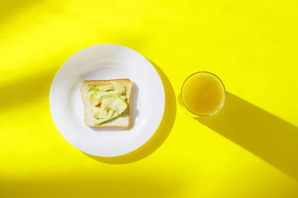 Σάντουιτς με αβοκάντο σε λευκό πιάτο και ένα ποτήρι με χυμό πορτοκαλιού σε κίτρινο φόντο. Υγιεινή αίσθηση πρωινού, βιταμίνες, δίαιτα, απώλεια βάρους. Επίπεδη όψη, κορυφή θέα. — Φωτογραφία Αρχείου