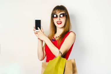 Kırmızı elbiseli genç bir kız, elinde telefon ve alışveriş çantaları yla hafif bir arka plan üzerinde. Online alışveriş konsepti, alışveriş uygulaması, satış.