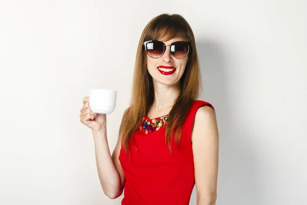 Una joven con un vestido rojo sostiene en su mano una taza de café sobre un fondo claro. concepto de cafetería, desayuno . — Foto de Stock