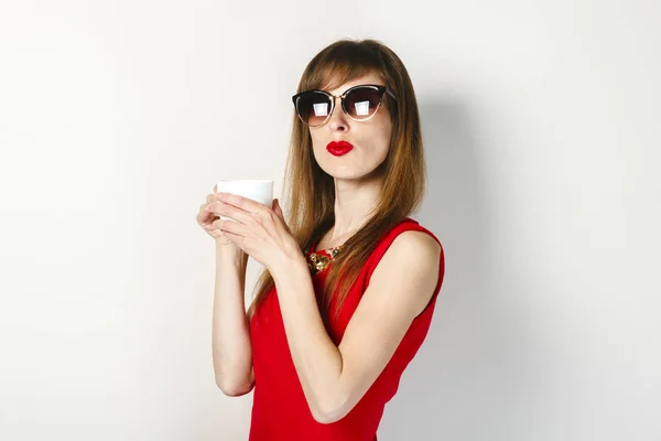 Una joven con un vestido rojo sostiene en su mano una taza de café sobre un fondo claro. concepto de cafetería, desayuno . — Foto de Stock