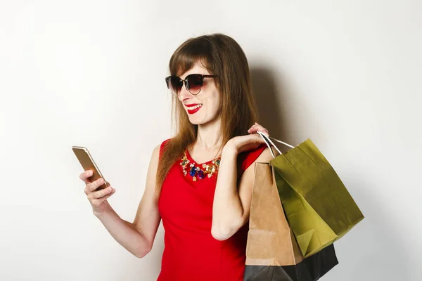 Uma menina em um vestido vermelho segurando um telefone e sacos de compras em um fundo leve. Conceito de compras on-line, aplicativo de compras, venda . — Fotografia de Stock