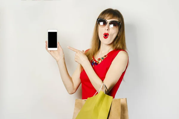 Młoda dziewczyna w czerwonej sukni trzyma telefon i torby na zakupy na jasnym tle. Koncepcja zakupów online, aplikacja na zakupy, sprzedaż. — Zdjęcie stockowe